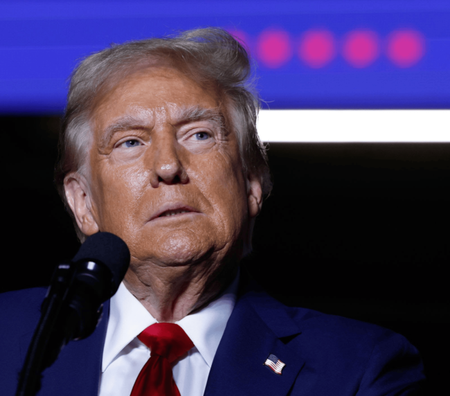 Close-up of President Trump, the elected 47th President of the USA in a suit and red tie speaking into a microphone, set against a background featuring blue and purple tones with blurred lights.