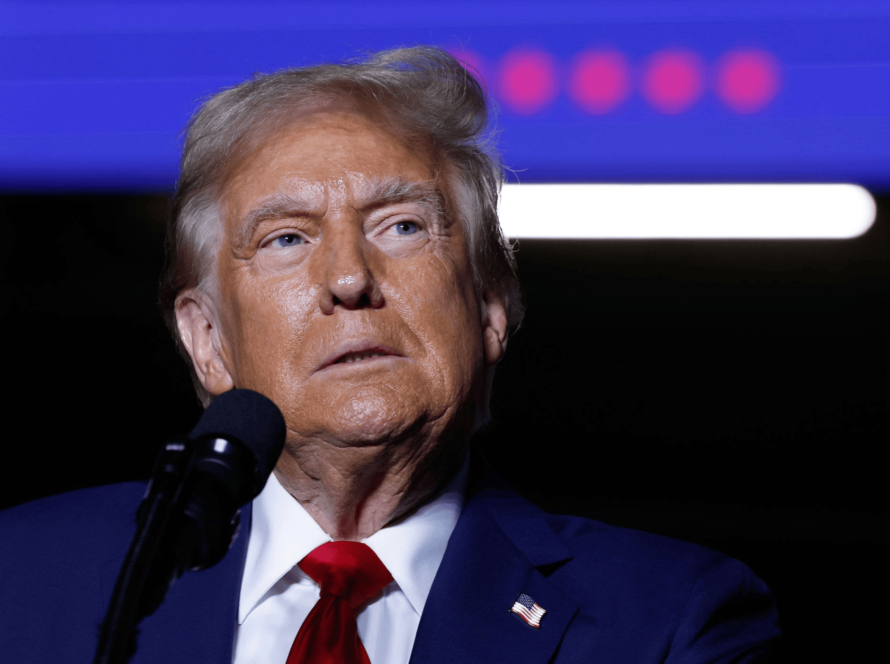 Close-up of President Trump, the elected 47th President of the USA in a suit and red tie speaking into a microphone, set against a background featuring blue and purple tones with blurred lights.